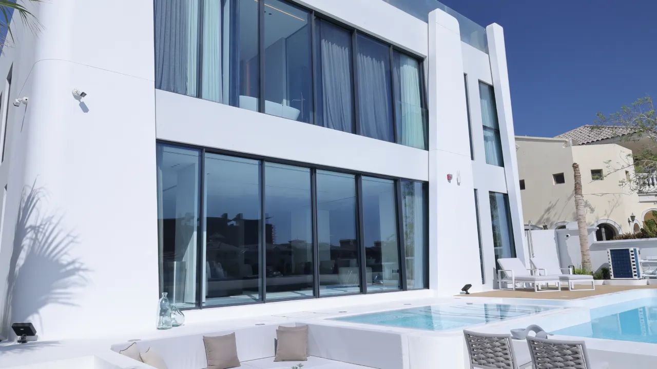 A modern white home exterior in Palm Jumeirah, Dubai, featuring large glass windows, a bright blue swimming pool, outdoor lounge chairs.