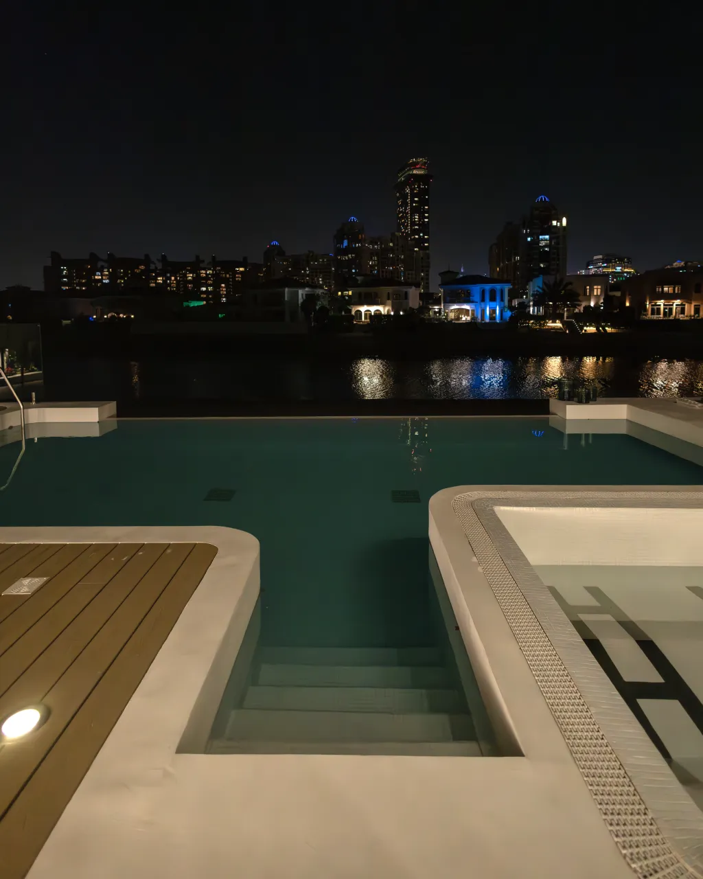 Night view of renovated waterfront swimming pool with dark water, canal reflections, city lights, and spa edge