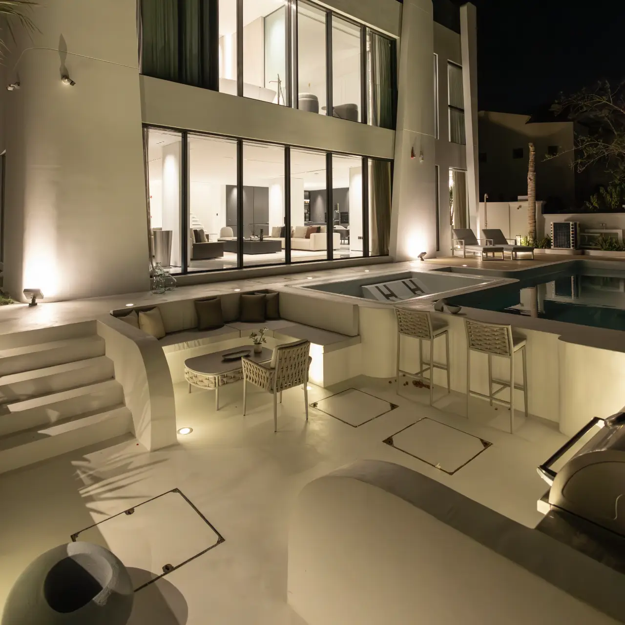 Night view of a luxury modern villa with large glass windows, warm exterior lighting, a swimming pool, bar stools, and a sunken outdoor lounge with cushioned seating.