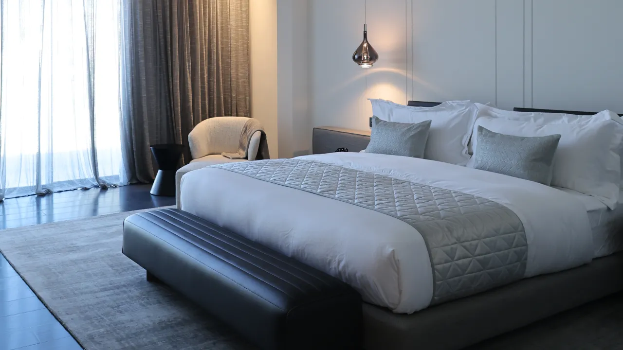 Luxury bedroom after villa renovation in Palm Jumeirah with layered white bedding, grey textiles, dark bench, and soft window drapery
