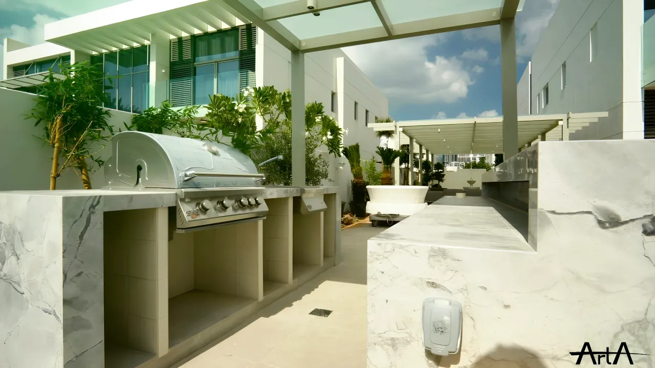 MBR District One modern villa side terrace with an BBQ kitchen, marble-look counters, built-in grill, open storage bays, pergola shade, and greenery along the wall.
