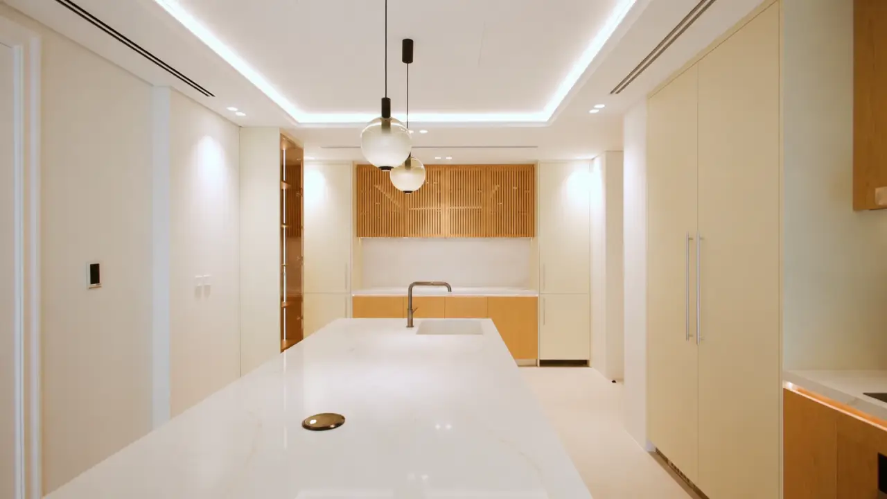 Kitchen renovation in Meadows Dubai with white island, pale wood cabinets, slatted upper storage, and globe pendant lights.