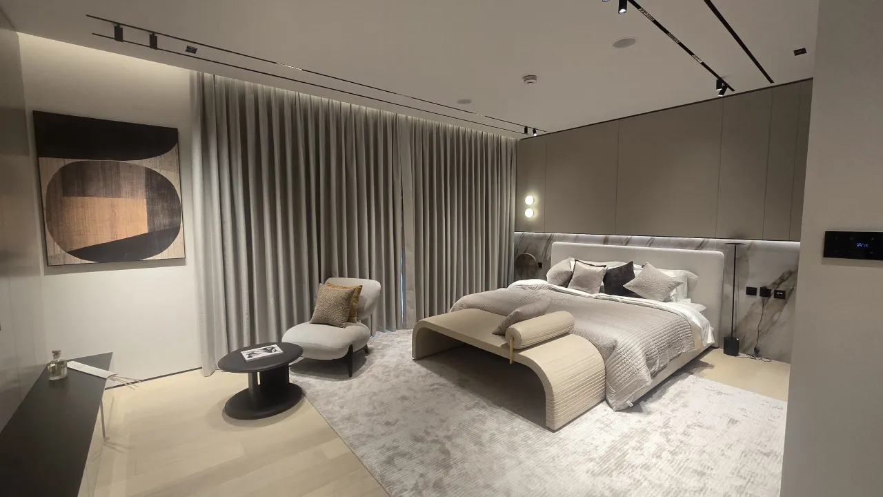 Modern bedroom suite renovation with soft grey curtains, upholstered bed, curved bench, lounge chair, black side table and warm wall lighting