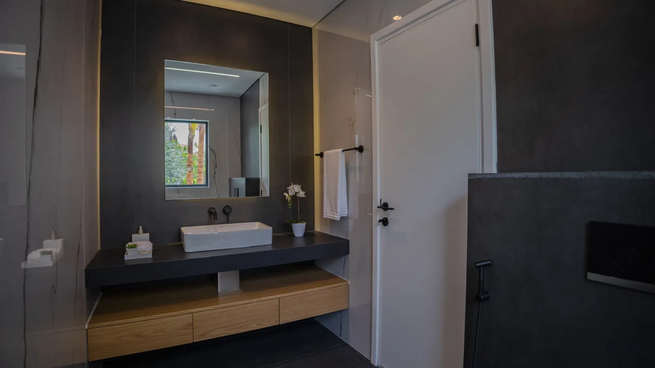 Modern villa bathroom renovation with black vanity wall, wood drawers, vessel sink, backlit mirror, marble-effect panels, and black fixtures