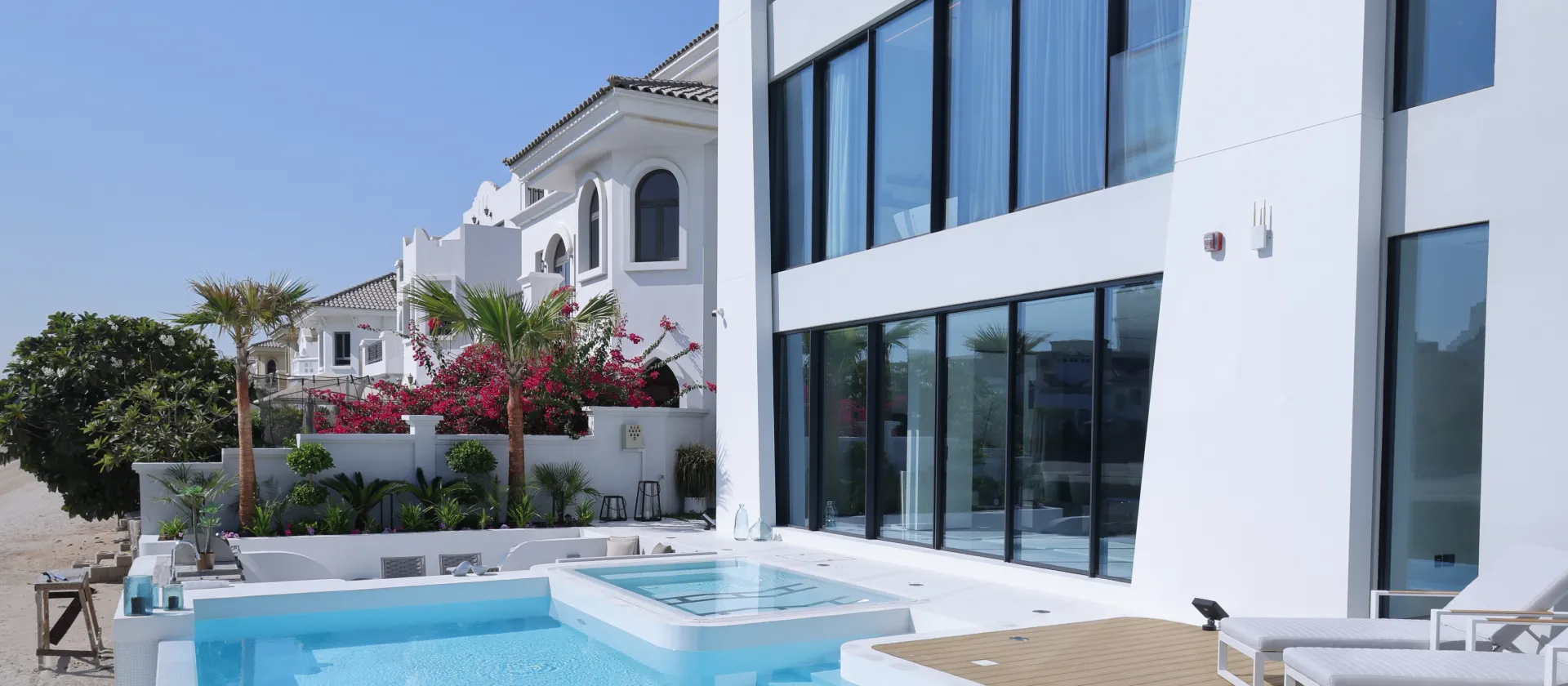 A modern white villa elevation in Palm Jumeirah with large black-framed glass windows, a bright swimming pool, sun loungers, palm trees, flowering landscaping, and neighboring luxury villas under a clear blue sky.