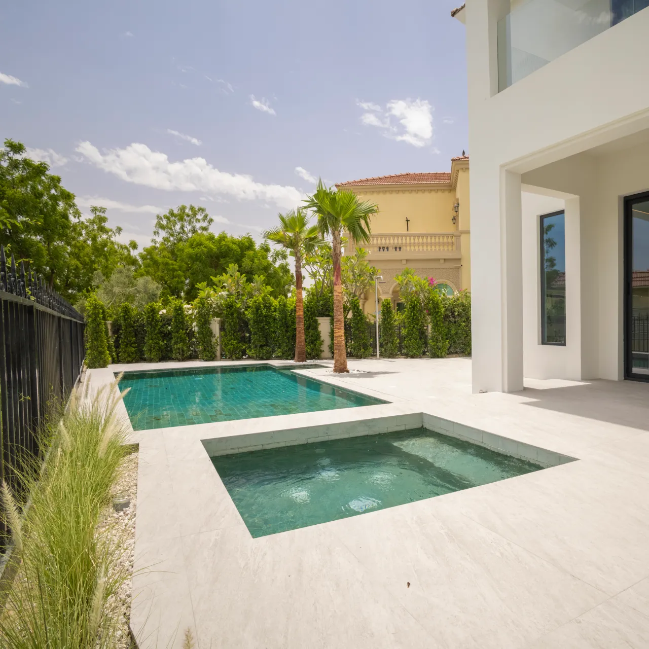 Private villa pool reconstruction with large rectangular pool with Jacuzzi, white villa facade, stone deck, palm trees, hedges, and garden privacy