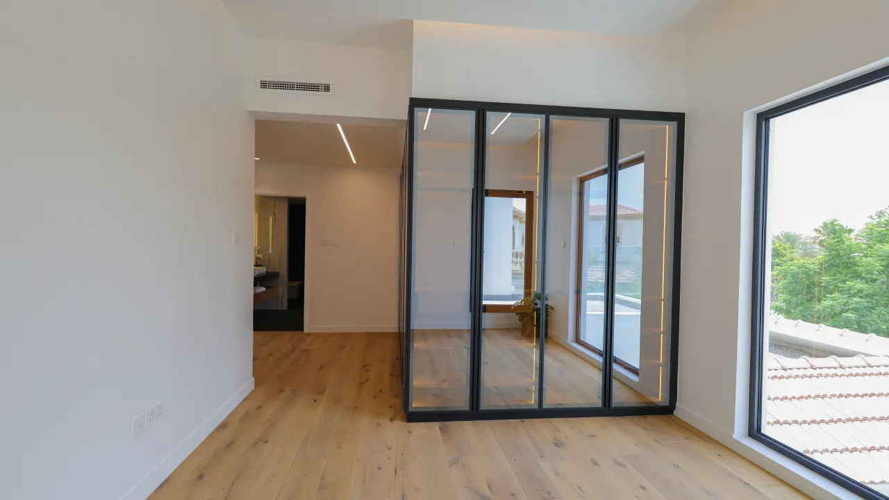 Renovated villa bedroom with warm wood floor, black-framed glass partition, white walls, large window, and bright modern interior