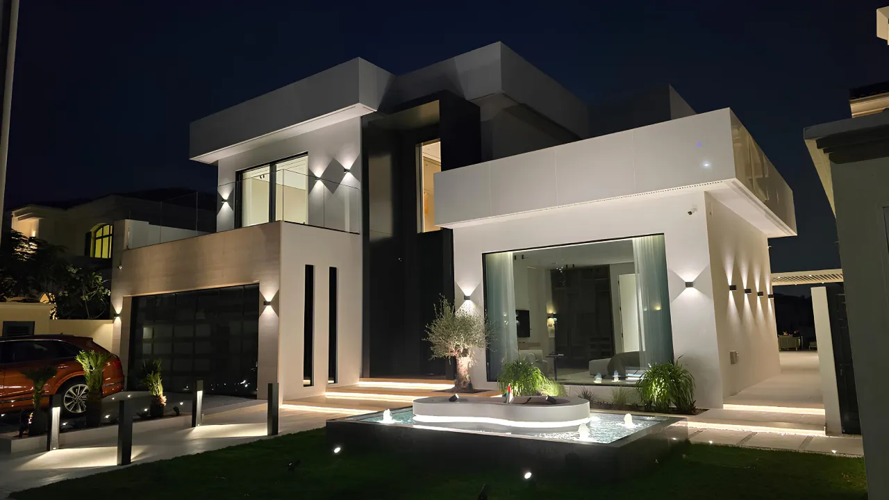 Contemporary luxury villa exterior at night with black-and-white facade, large windows, warm wall lights, landscaped lawn, and front fountain garden.