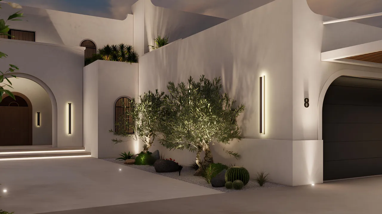 White villa entrance at night with arched doorway, driveway, warm wall lights, black garage door, palms, and layered garden planting.