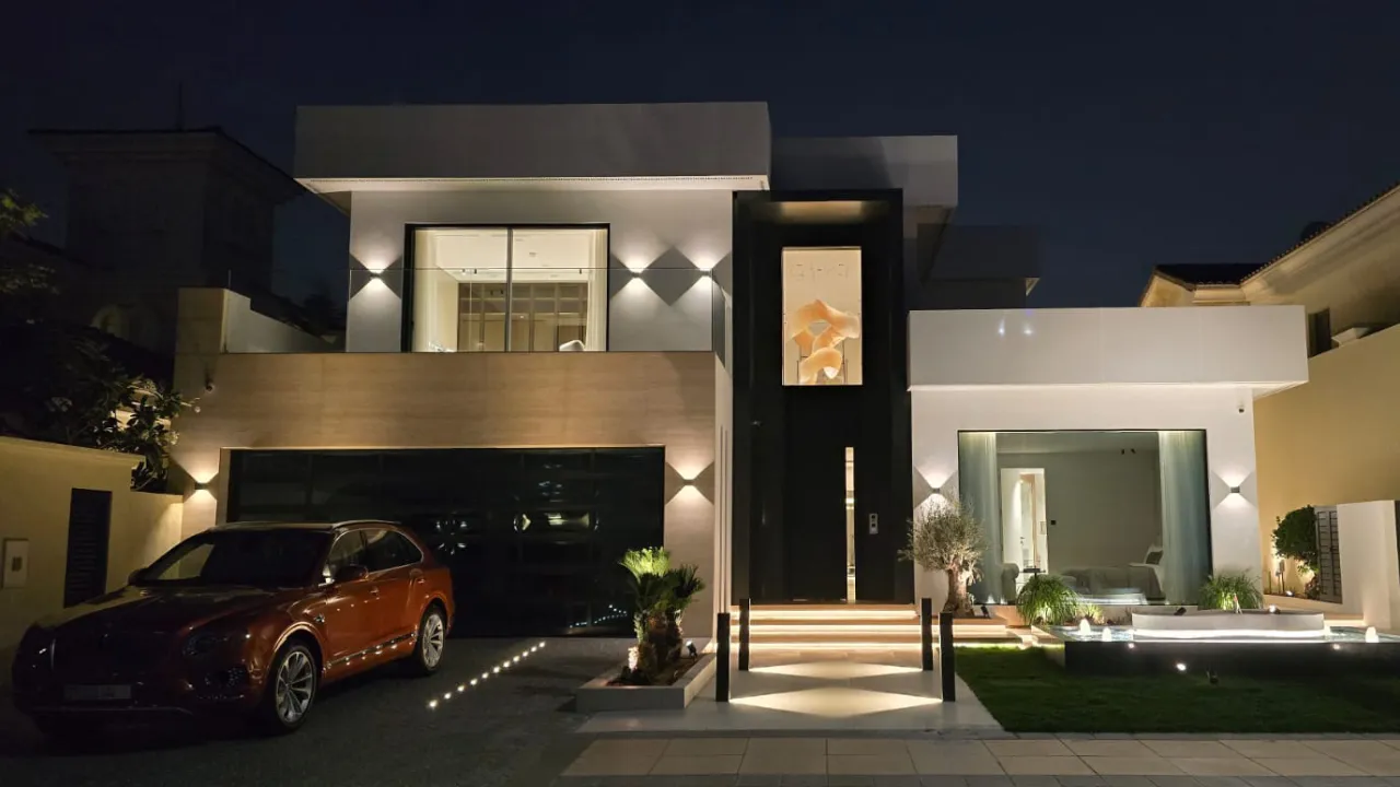 Modern Palm Jumeirah villa entrance with driveway hard landscaping, paved path, garden borders, warm lighting, glass facade, and structured front garden.