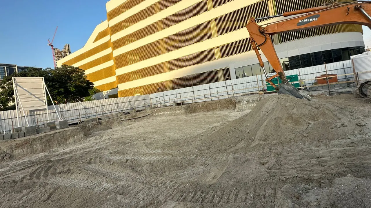 Excavator working on a Dubai construction site with soil excavation, site fencing, and a large yellow-and-white building in the background.