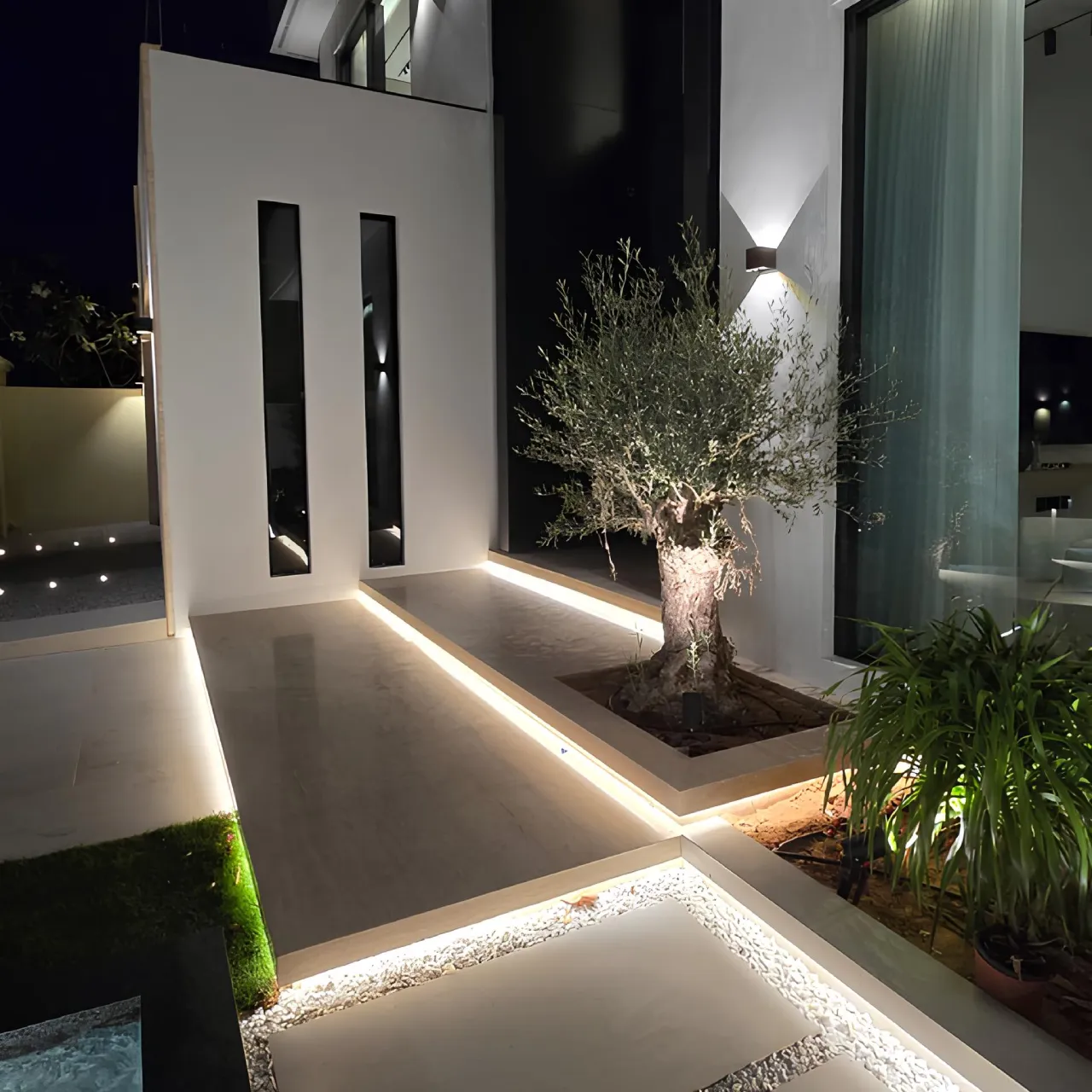 A modern house entrance after renovation at night, featuring a white and black facade, slim vertical windows, warm linear pathway lighting, an illuminated olive tree, planted borders, and a minimalist walkway.