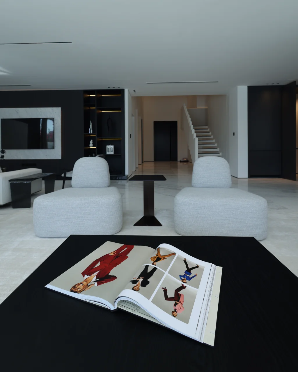 Close-up lounge view with black coffee table, open book styling, rounded chairs, marble floor, dark TV wall, display shelving, and staircase background