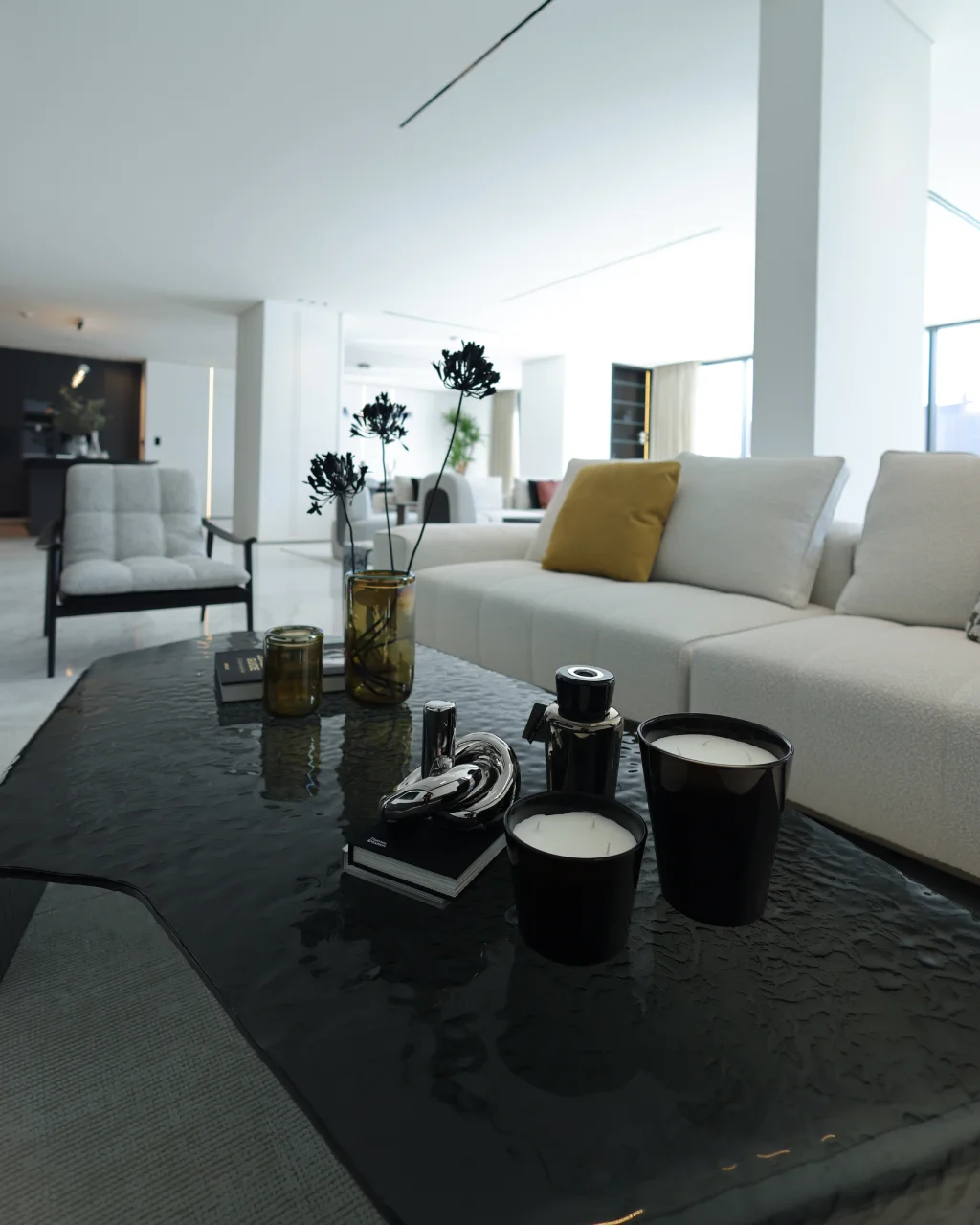 Close-up of glossy black coffee table with candles, amber glass vases, silver decor, cream sofa, mustard pillow, and open-plan villa background