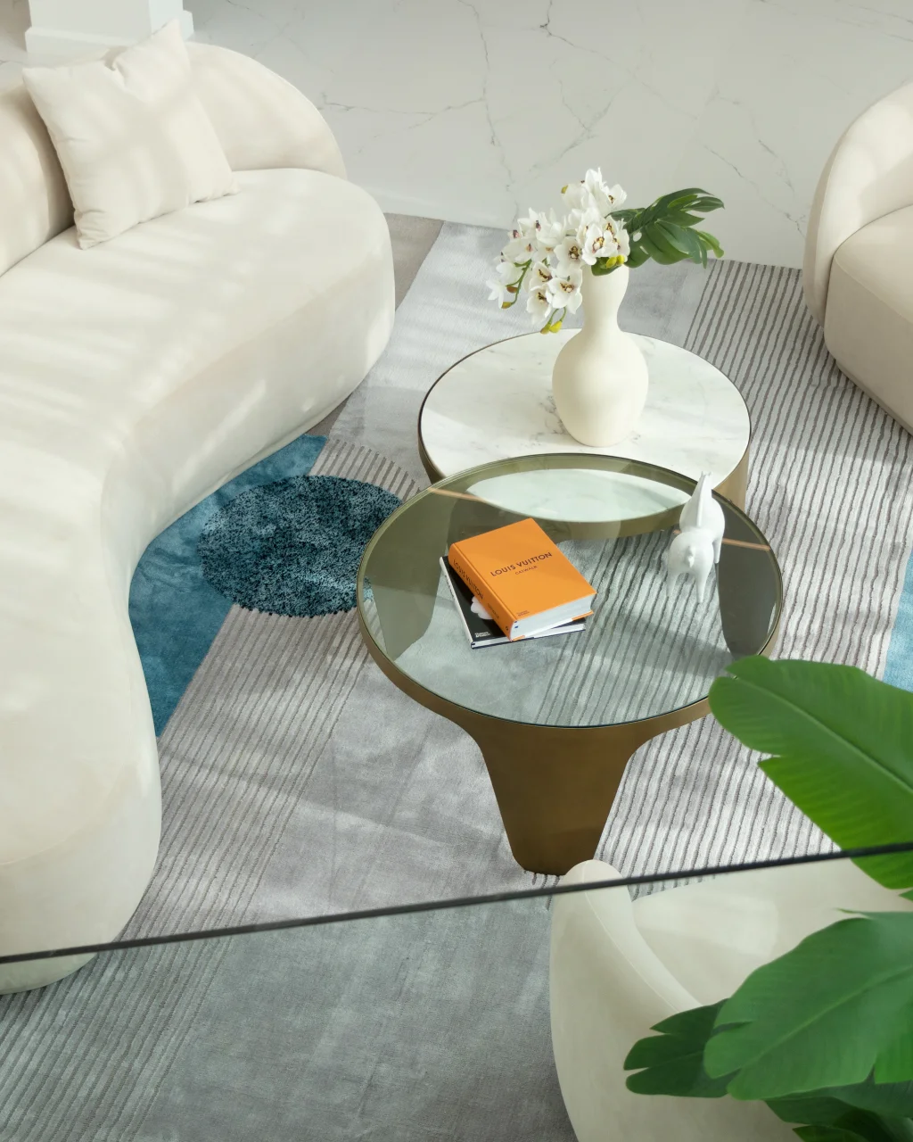 Close-up lounge styling with glass coffee table, white orchids, orange coffee table book, cream curved seating, blue accent rug, and marble floor