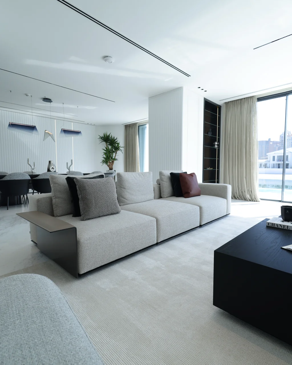 Contemporary waterfront sofa area with light textured sofa, black coffee table, open dining room, vertical wall paneling, beige curtains, and marble flooring