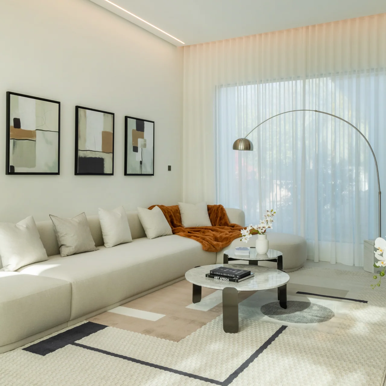 Cream family lounge with abstract wall art, curved sectional sofa, rust throw, layered marble coffee tables, arched floor lamp, and geometric rug
