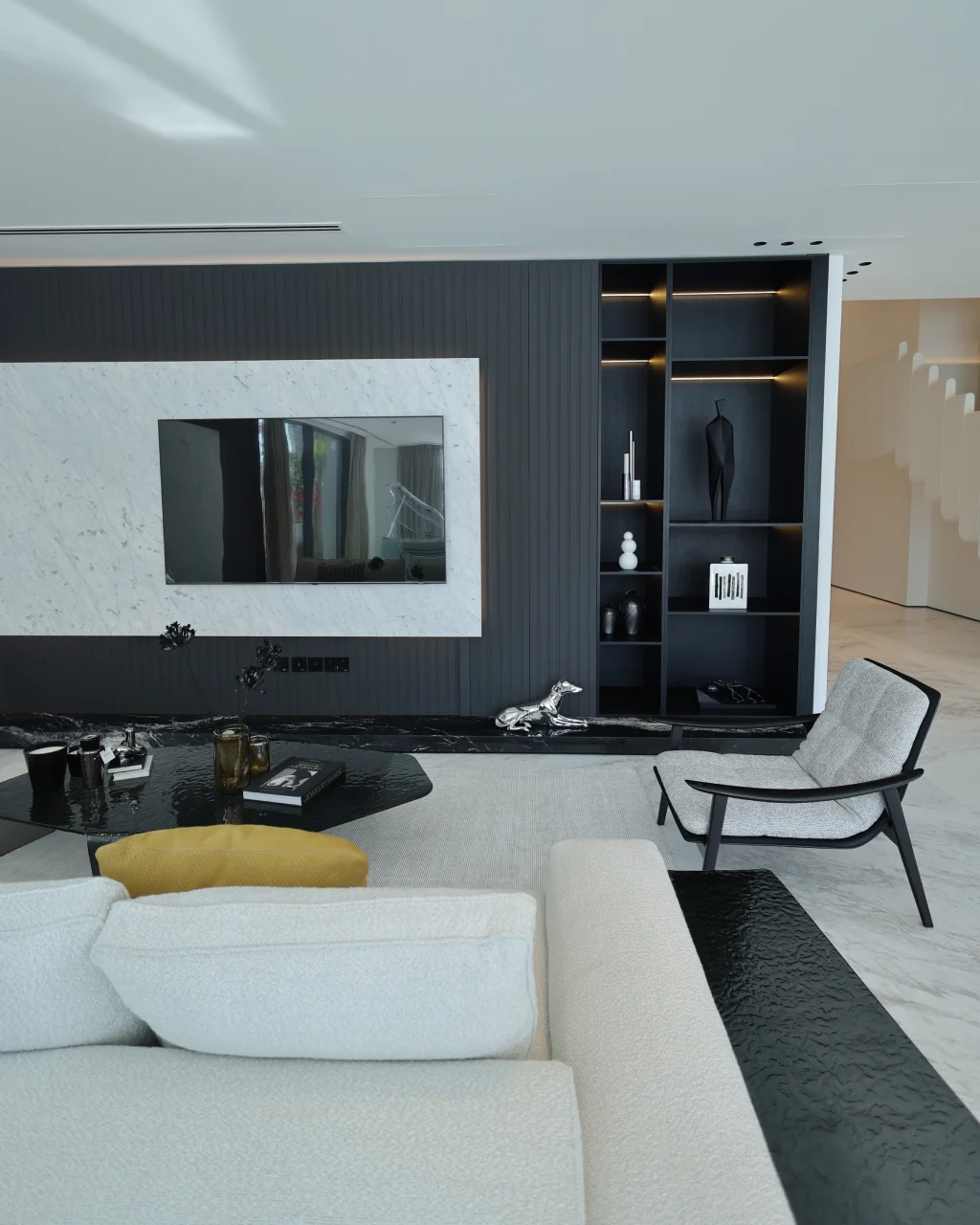 Dark media wall living room with illuminated black shelving, marble TV surround, cream sofa, black stone ledge, slim lounge chair, and staircase view