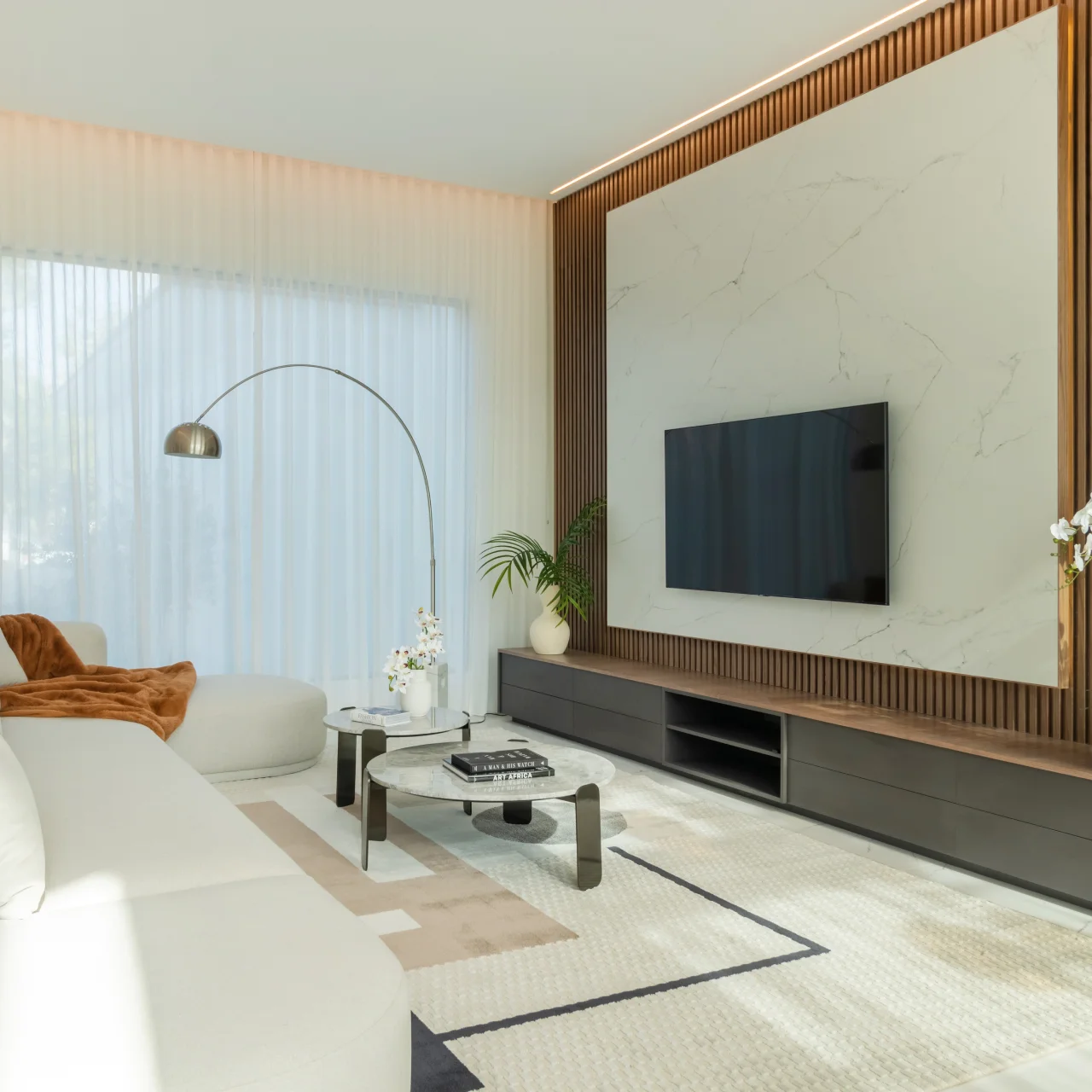 Jumeirah Islands family living room with marble TV wall, warm wood slat paneling, cream sofa, arched floor lamp, round coffee tables, and sheer curtains