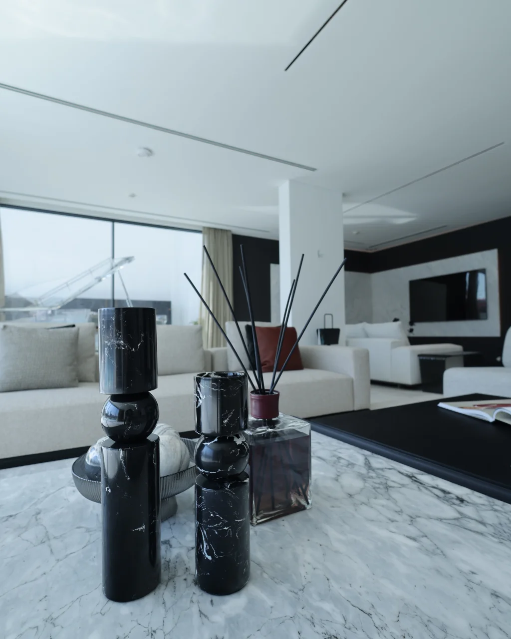Marble coffee table detail with glossy black decorative objects, reed diffuser, cream sofa, dark TV wall, pale marble floor, and monochrome living room palette
