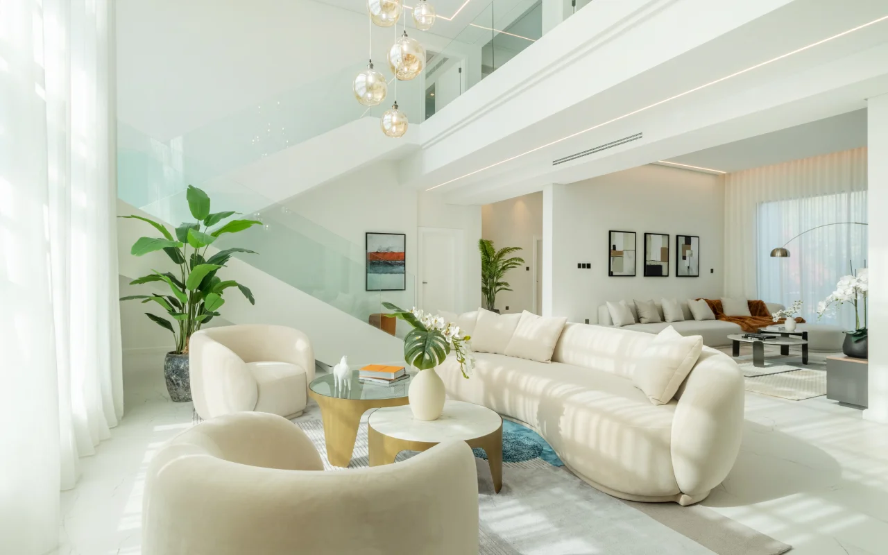Open double-height villa lounge with curved cream sofa, indoor plants, glass railing staircase, amber pendants, blue accent rug, and secondary living area