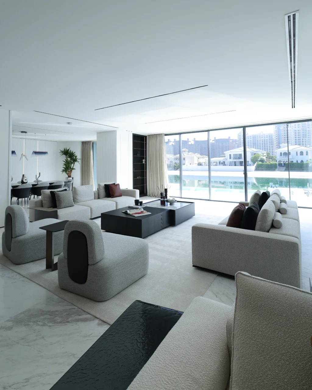 Palm Jumeirah living room with waterfront view, cream sofas, rounded lounge chairs, black coffee table, open dining area, marble floor, and beige curtains