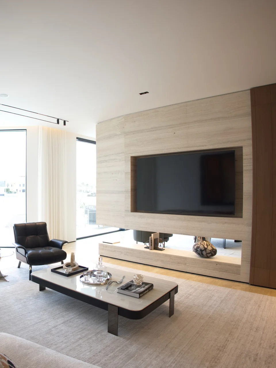 Palm Jumeirah TV lounge with beige stone media wall, recessed television, warm wood paneling, black leather chair, rectangular coffee table, and sheer curtains