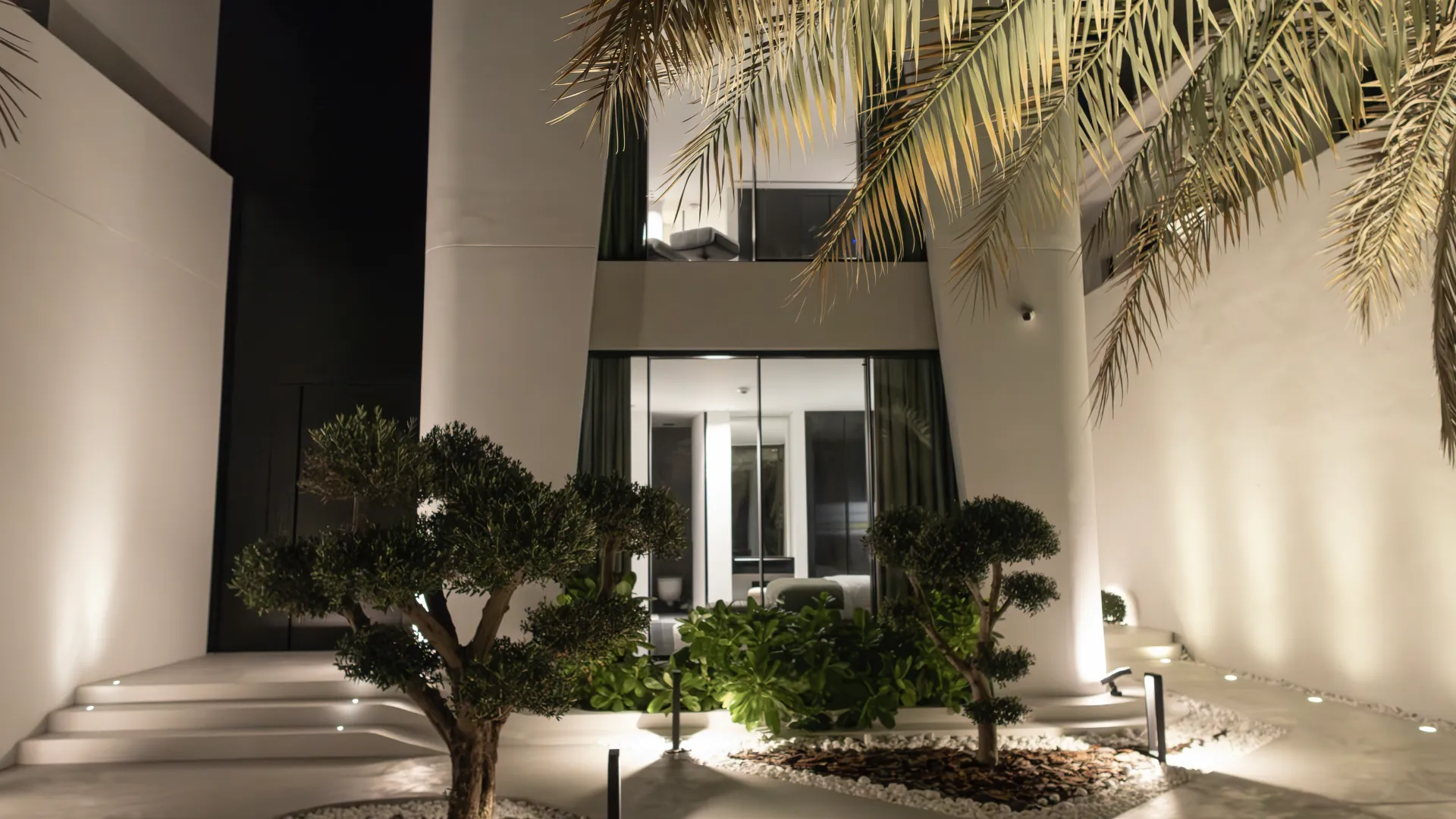Night view of the remodeled Palm Jumeirah villa entrance with illuminated landscaping, sculptural trees, curved steps and tall black-framed glazing.