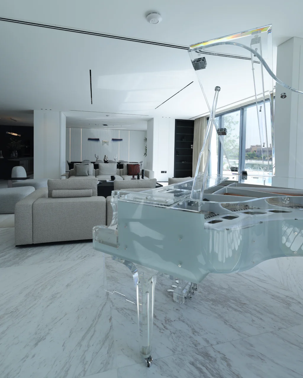 Transparent grand piano in Palm Jumeirah living room with cream seating, black coffee table, open dining area, marble flooring, and waterfront daylight