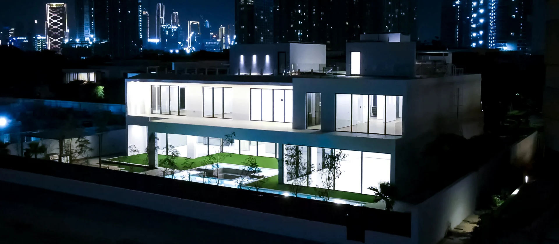 Night aerial view of luxury custom house in Sobha Hartland Estates showing the illuminated villa, pool courtyard and Dubai skyline
