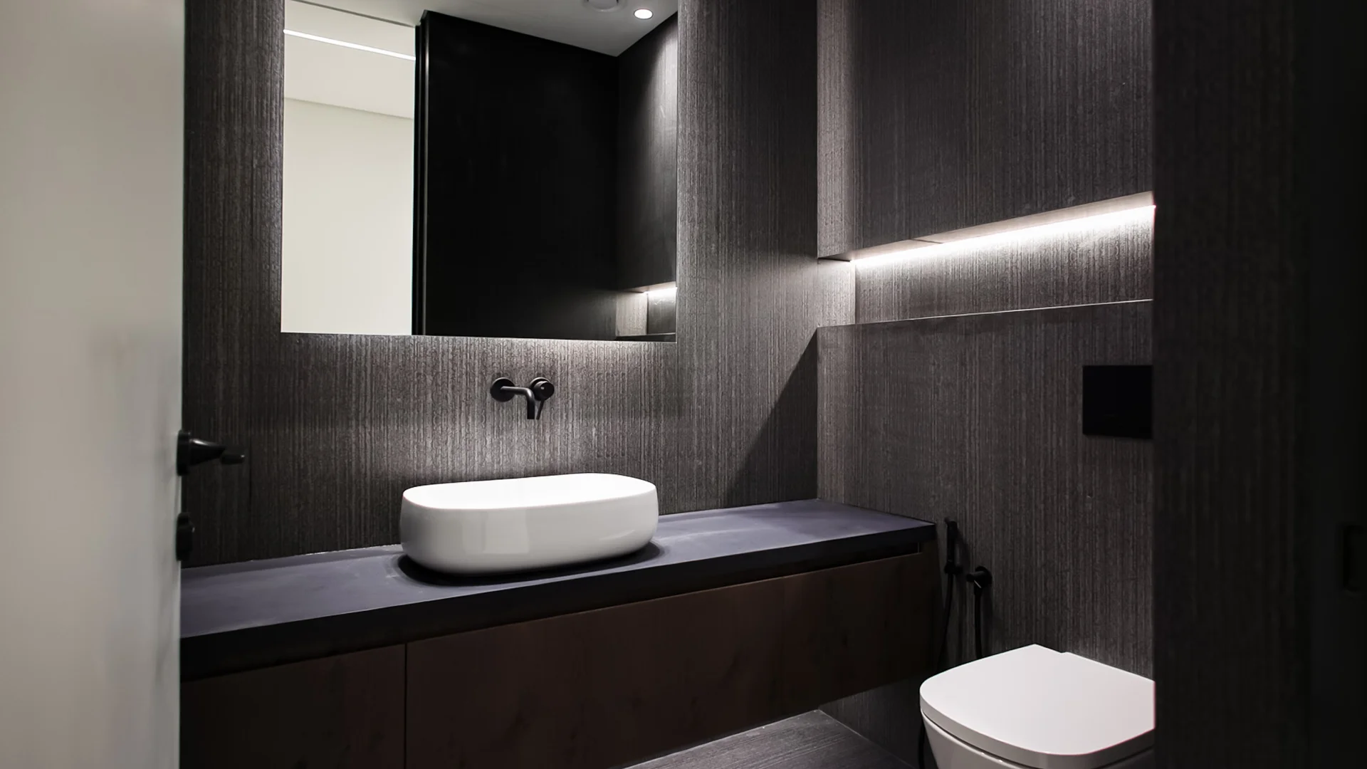 Dark powder room with floating timber vanity, white vessel basin, wall-mounted tap and concealed lighting